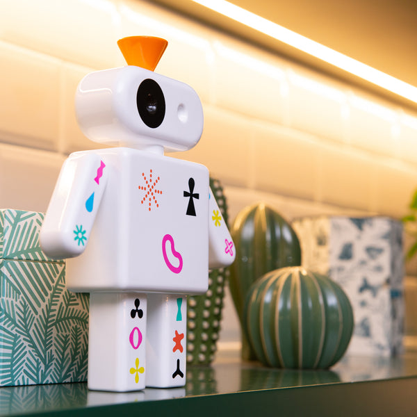 Ein kleiner weißer Spielzeugroboter mit bunten Symbolen steht auf einem Regal neben gemusterten Blumentöpfen.