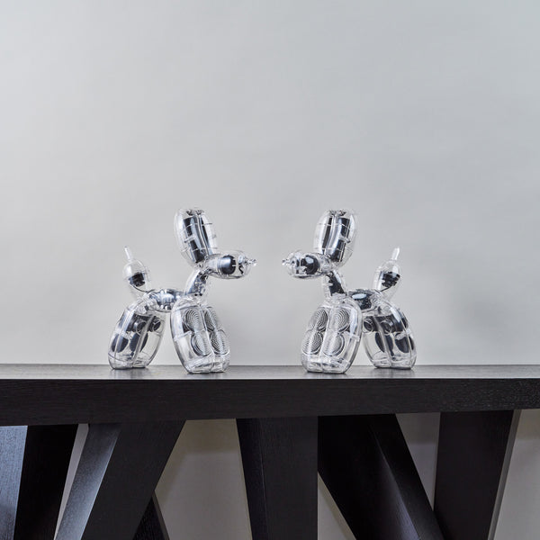 Zwei silberne Ballonhundeskulpturen auf einem dunklen Holztisch vor einem schlichten hellen Hintergrund.