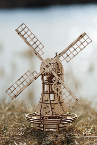 Ein Holzmodell einer Windmühle mit detaillierten Zahnrädern und Flügeln wird im Freien auf trockenem Gras in der Nähe von Wasser aufgestellt.