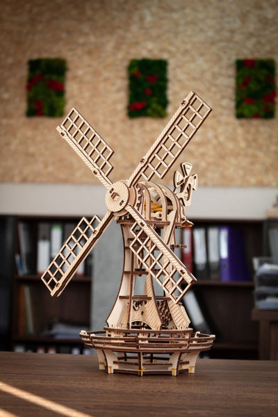 Ein Holzmodell einer Windmühle mit beweglichen Teilen steht auf einem Tisch in einer Büroumgebung mit Bücherregalen und Wanddekorationen im Hintergrund.