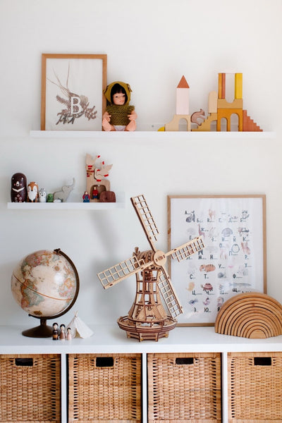 Kinderzimmerregale mit Spielzeug, Büchern, einem Globus, einer hölzernen Windmühle, Weidenkörben, Tierfiguren und gerahmter Kunst vor einer weißen Wand.
