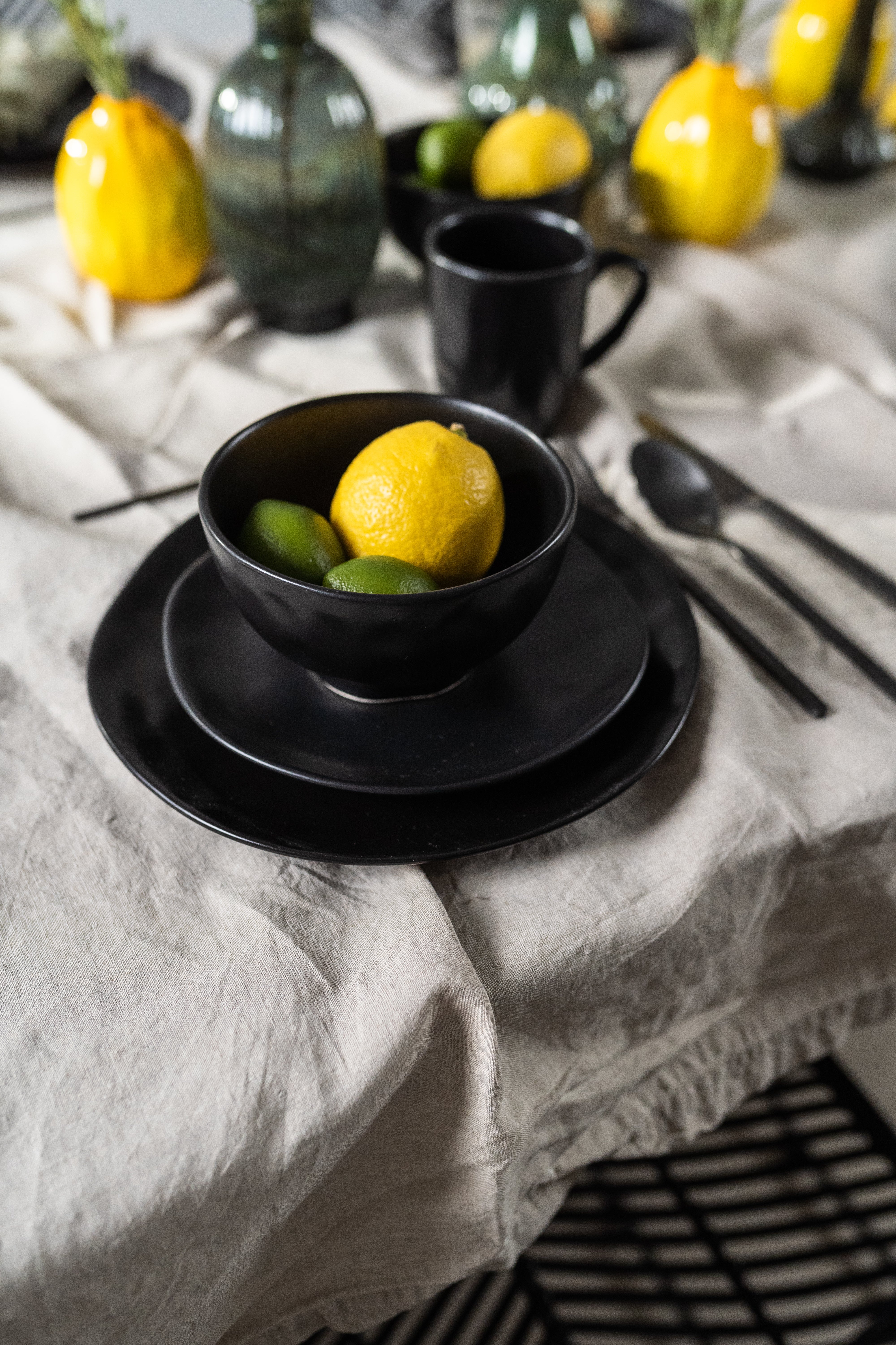 Eine Schale „Organic Black“ (Ø 15 cm) mit einer Zitrone und zwei Limetten steht auf einem passenden Teller neben schwarzem Besteck auf einer beigen Tischdecke, während grüne Vasen und gelbe Zitronen das Keramikgeschirr in handgefertigter Optik hervorheben.