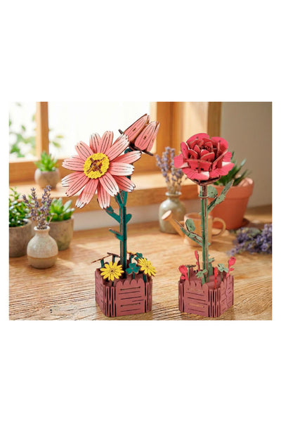 Zwei bunte, blockartige Modelle eines rosa Gänseblümchens und einer roten Rose mit grünen Stielen und Blättern stehen auf einem Holztisch in der Nähe eines Fensters mit Topfpflanzen im Hintergrund.