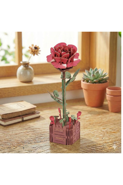 Eine Rosenskulptur aus Holz mit grünem Stiel und Blättern steht auf einem Holztisch neben Topfpflanzen, einer Vase und geschlossenen Büchern an einem Fenster.