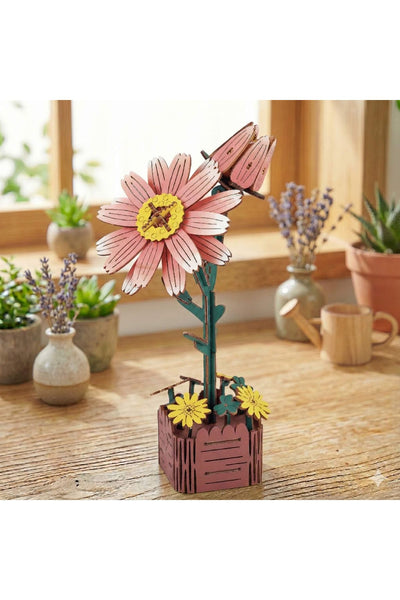 Eine Holzskulptur eines rosa-gelben Gänseblümchens mit zwei Knospen in einem kleinen kastenförmigen Topf steht auf einem Holztisch, umgeben von Topfpflanzen und Keramikgefäßen.