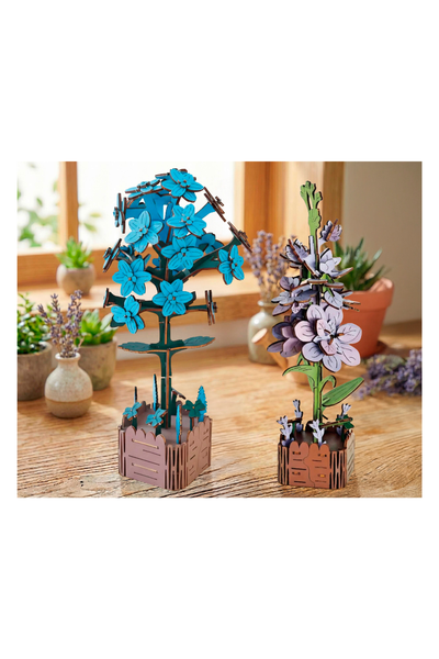 Zwei hölzerne Modellblumenskulpturen, eine mit blauen und eine mit violetten Blumen, stehen auf einem Holztisch vor einem Fenster mit Topfpflanzen im Hintergrund.