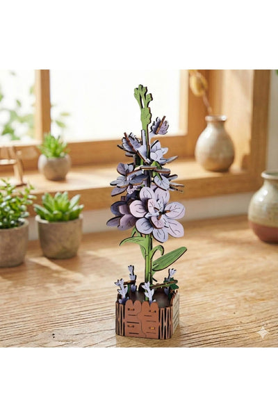 Ein hölzernes 3D-Puzzle-Modell mit lila Blumen in einem kleinen dekorativen Topf wird auf einem Holztisch ausgestellt, mit Topfpflanzen und Keramikvasen im Hintergrund.