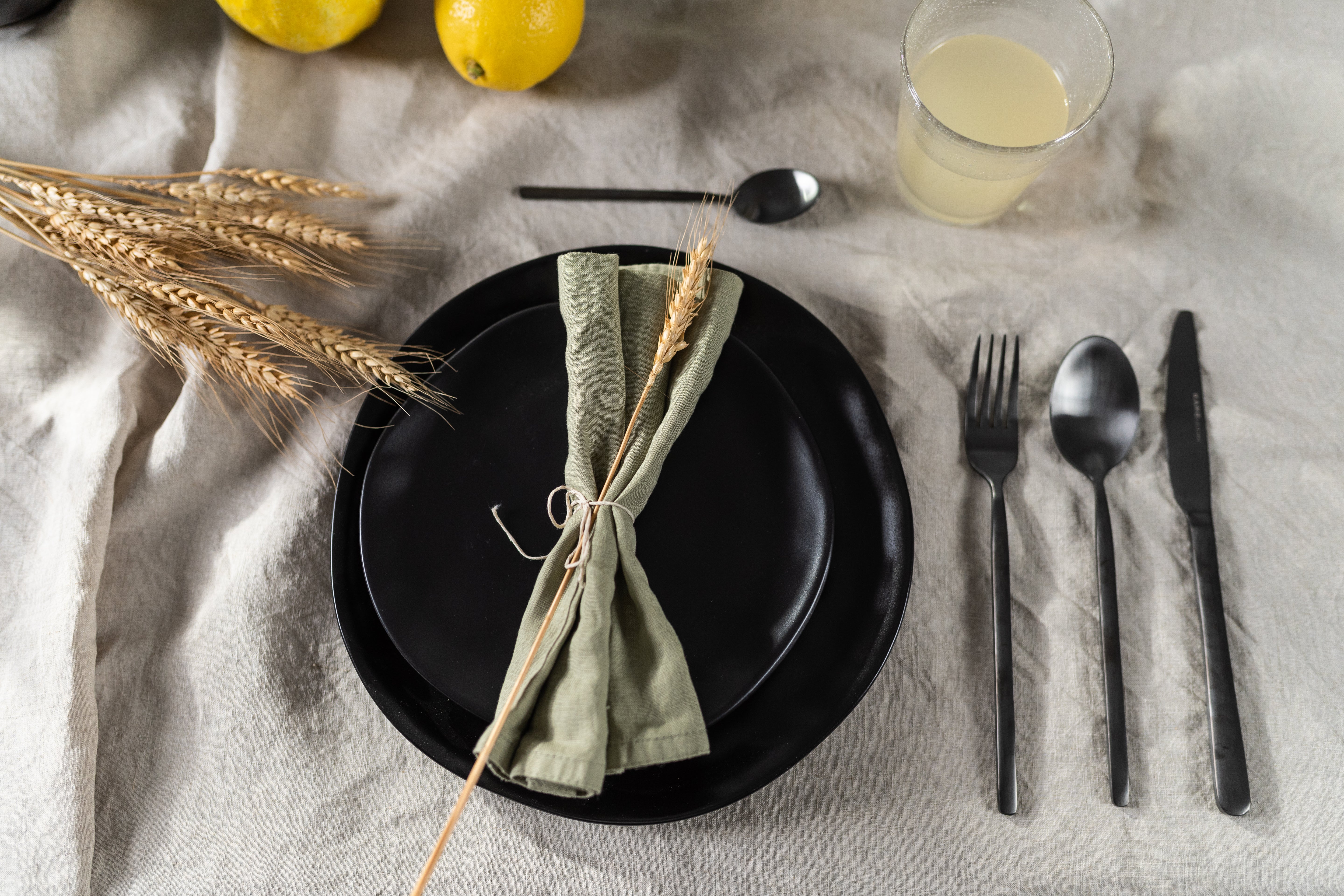Ein gedeckter Tisch mit dem Teller „Teller Organic Black“ (Ø 26 cm), einer grünen, mit Schnur und Weizen zusammengebundenen Serviette, schwarzem Besteck auf einer beigen Tischdecke sowie einem Glas hellem Saft und Zitronen in der Nähe.