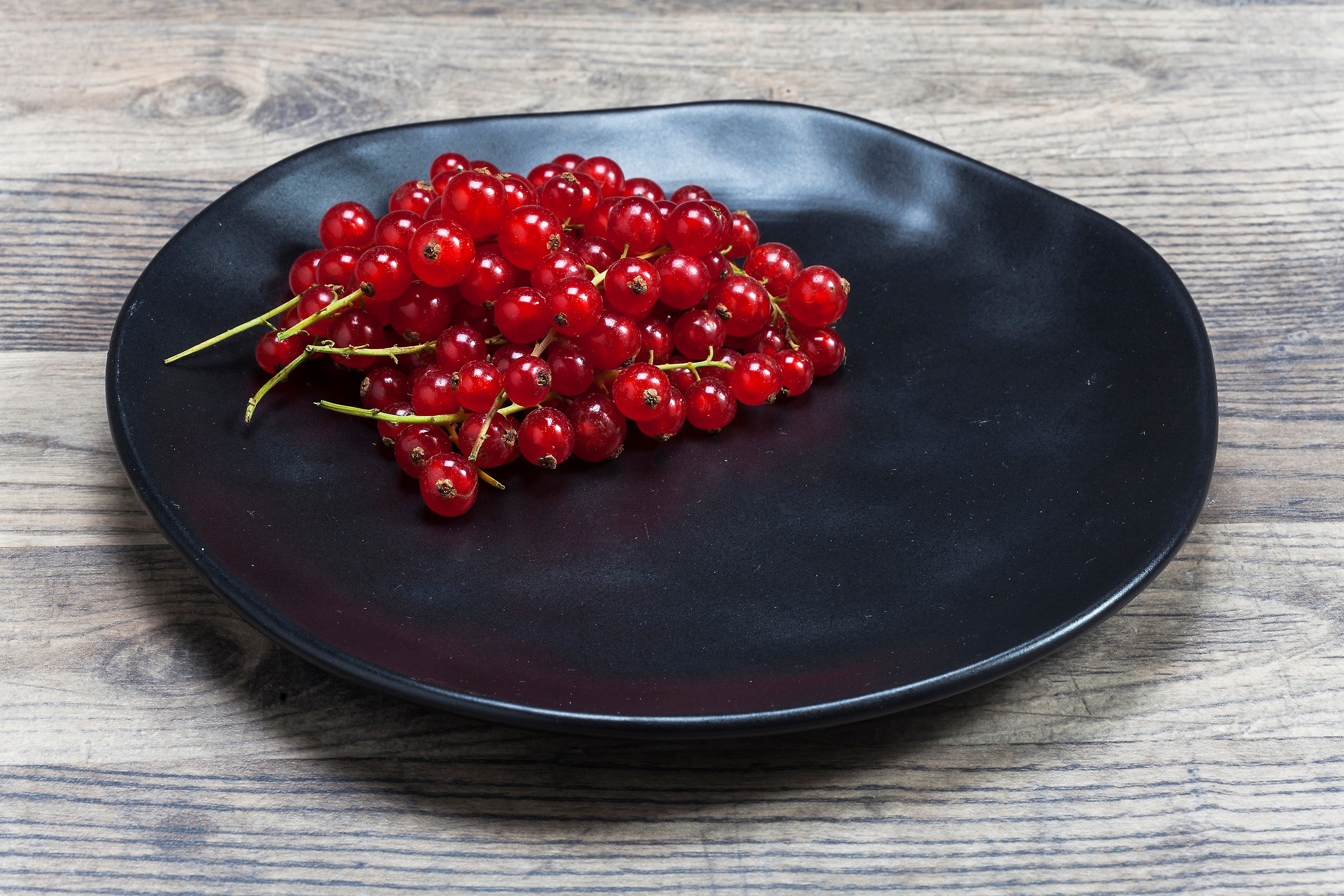 Eine Traube leuchtend roter Johannisbeeren liegt auf einem Teller Organic Black mit 20 cm Durchmesser, der auf einer rustikalen Holzoberfläche ruht.