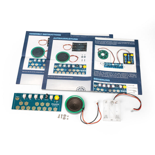 Elektronischer Bausatz mit Platine, Lautsprecher, Batteriehalter, Schrauben und Anleitungsblättern auf einer weißen Fläche.