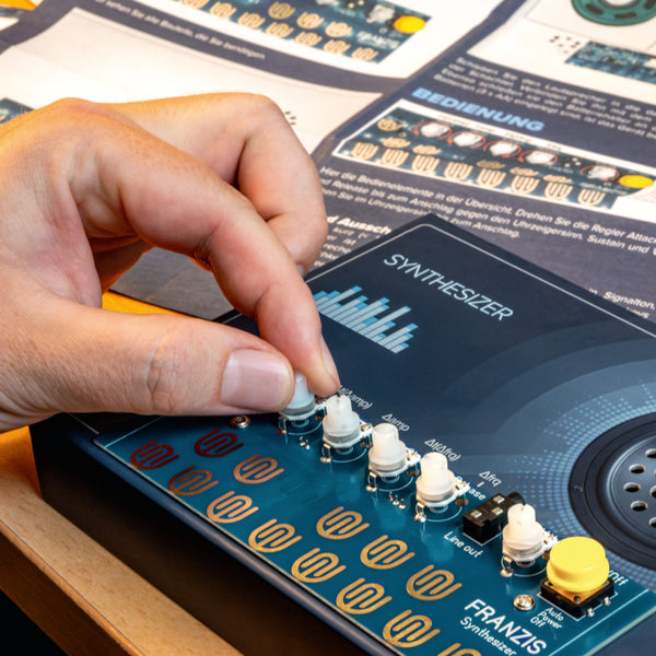 Eine Hand stellt einen Knopf an einem blauen Synthesizer ein, in dessen Nähe sich beschriftete Bedienelemente und Anleitungsblätter befinden.