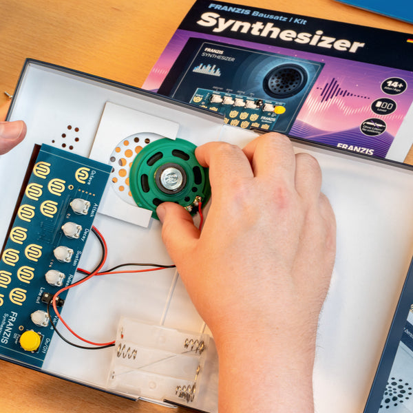 Hände beim Zusammenbau eines Synthesizer-Bausatzes mit Leiterplatten und Drähten, im Hintergrund ist die Bedienungsanleitung zu sehen.