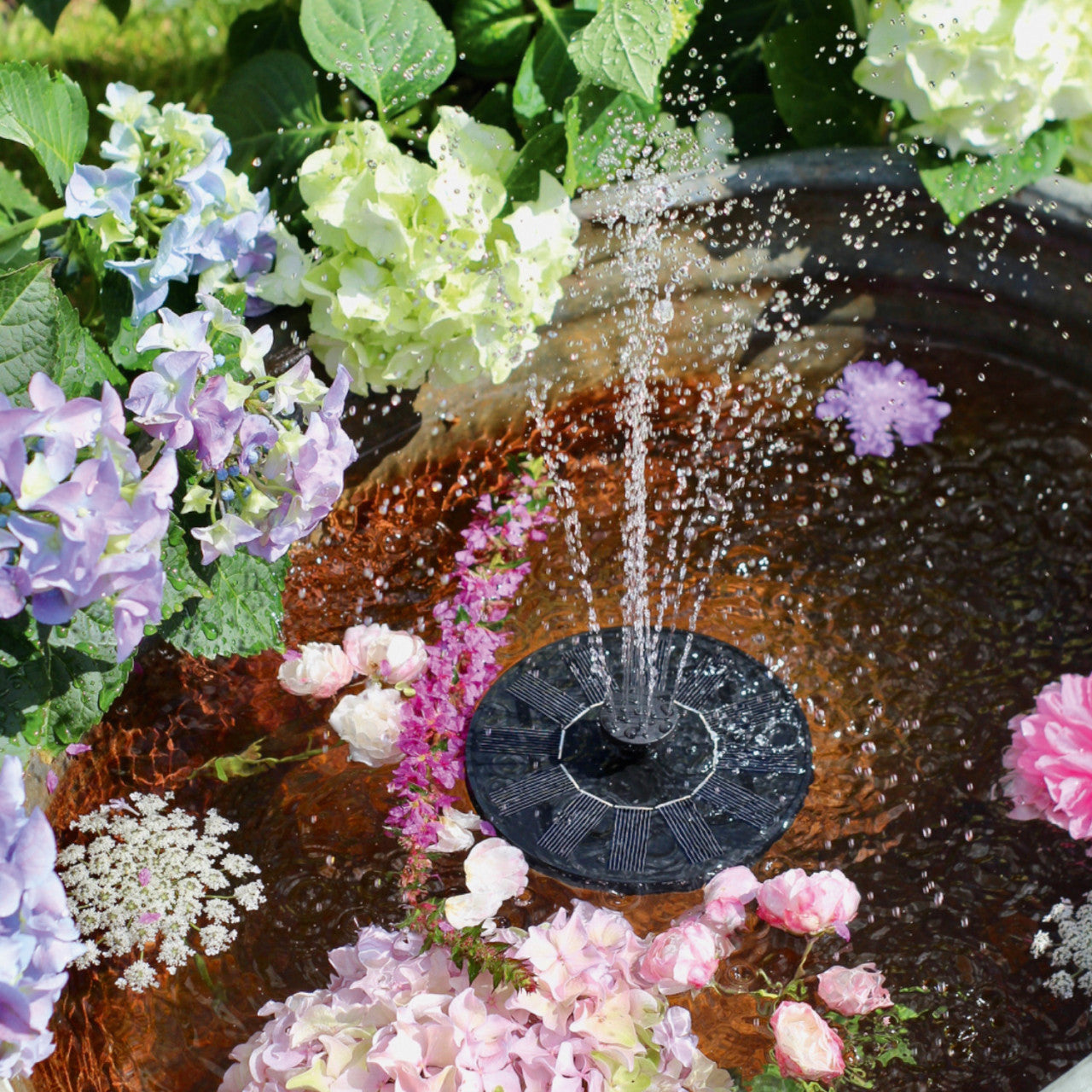 Der Blatt & Blüte Solar-Brunnen sprudelt sanft Wasser in eine Schale, umgeben von leuchtenden Hortensien und anderen bunten Blumen.