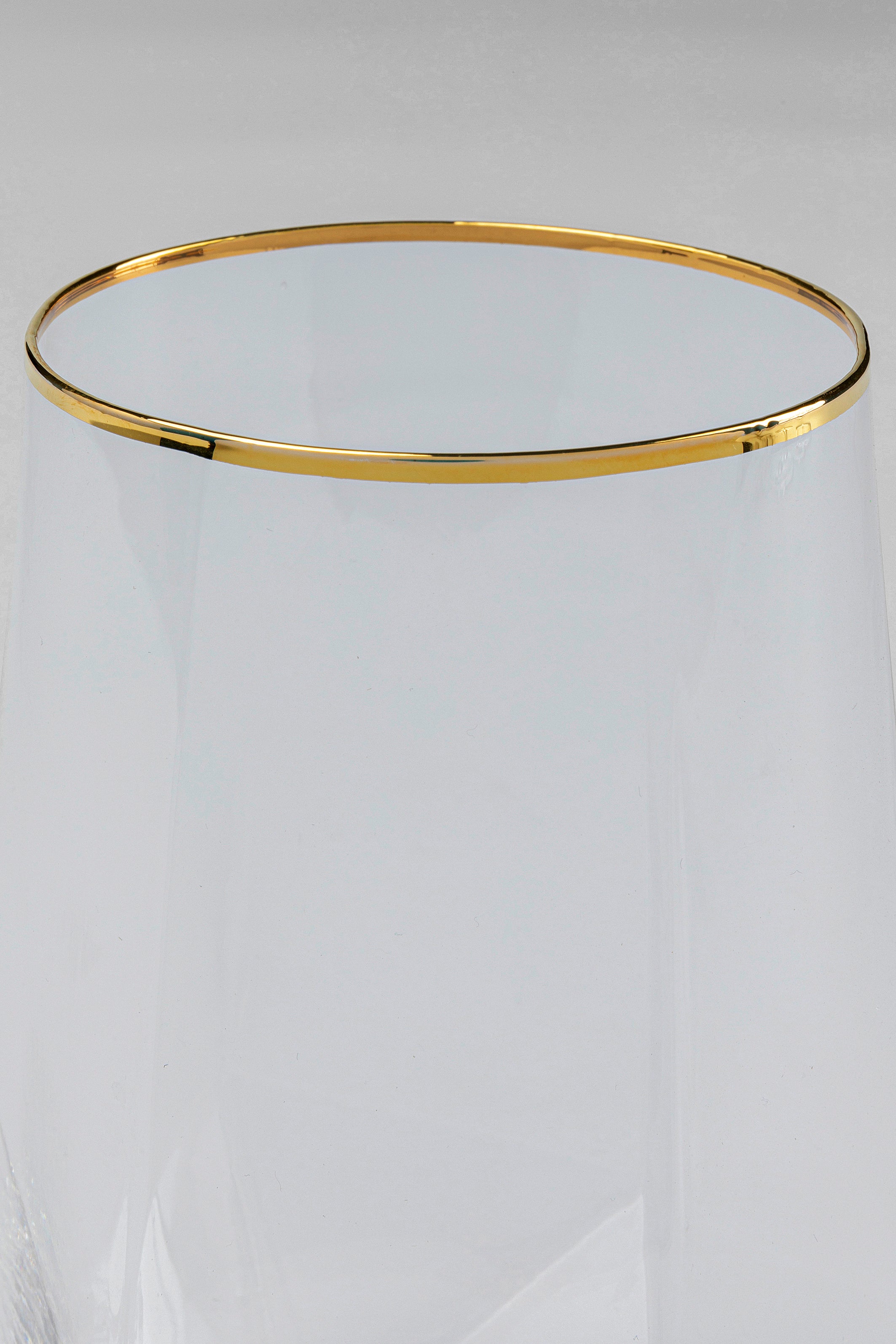 Eine Nahaufnahme des Randes eines Wasserglases Diamond Gold Rim (4/tlg.) mit einem dünnen Goldrand vor einem schlichten, hellgrauen Hintergrund.