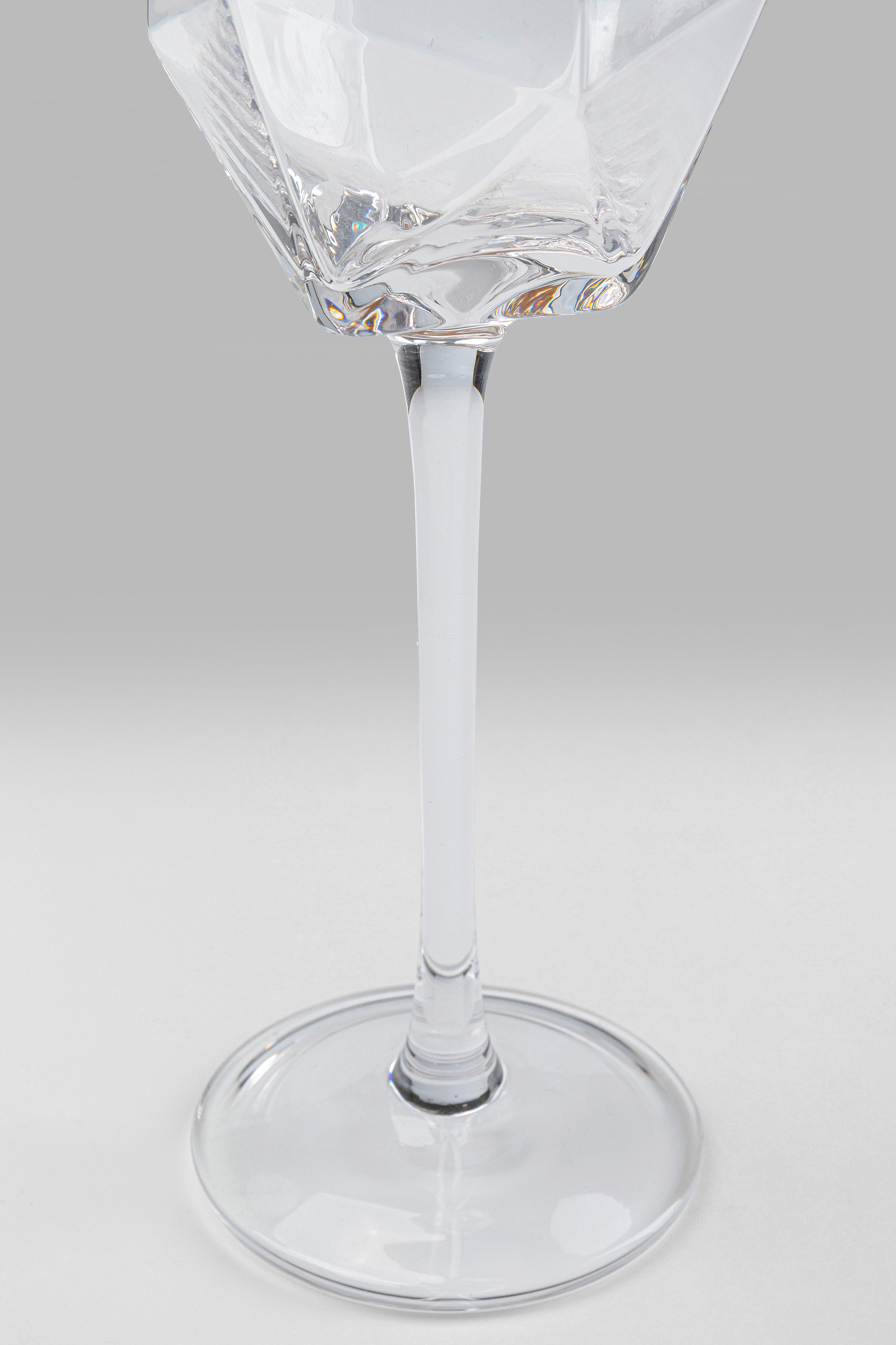 Eine Nahaufnahme des Sektglases Diamond Gold Rim (4/tlg.), ein klares Weinglas mit geometrischem, diamantinspiriertem Design und langem Stiel, präsentiert auf einer flachen Oberfläche vor einem hellgrauen Hintergrund.