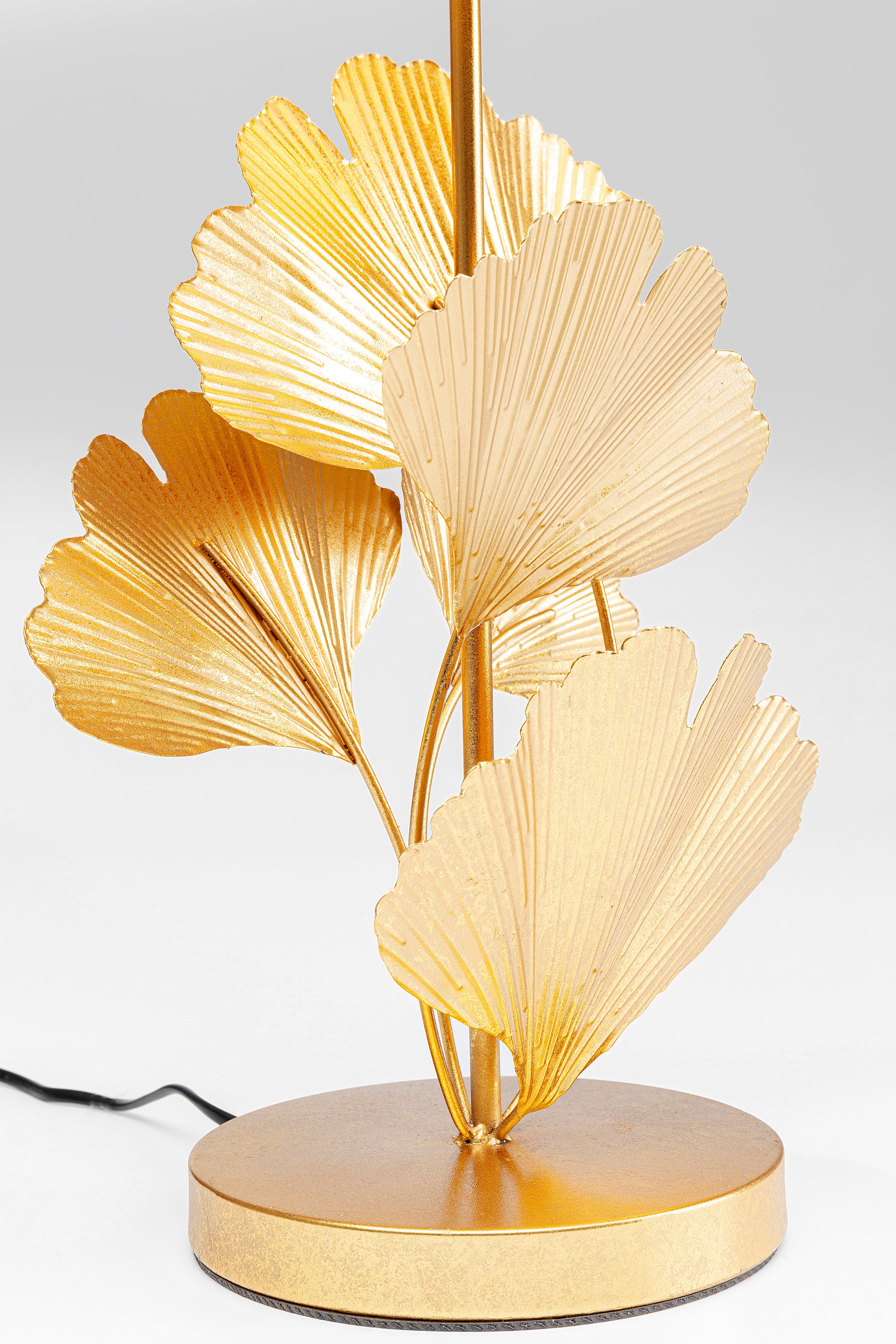 Eine Nahaufnahme der Tischleuchte Flores Gold, einer stilvollen Dekolampe mit goldfarbenen Ginkgoblättern aus Metall, rundem Sockel, sichtbarem schwarzen Netzkabel und schickem asiatischen Flair vor hellem Hintergrund.