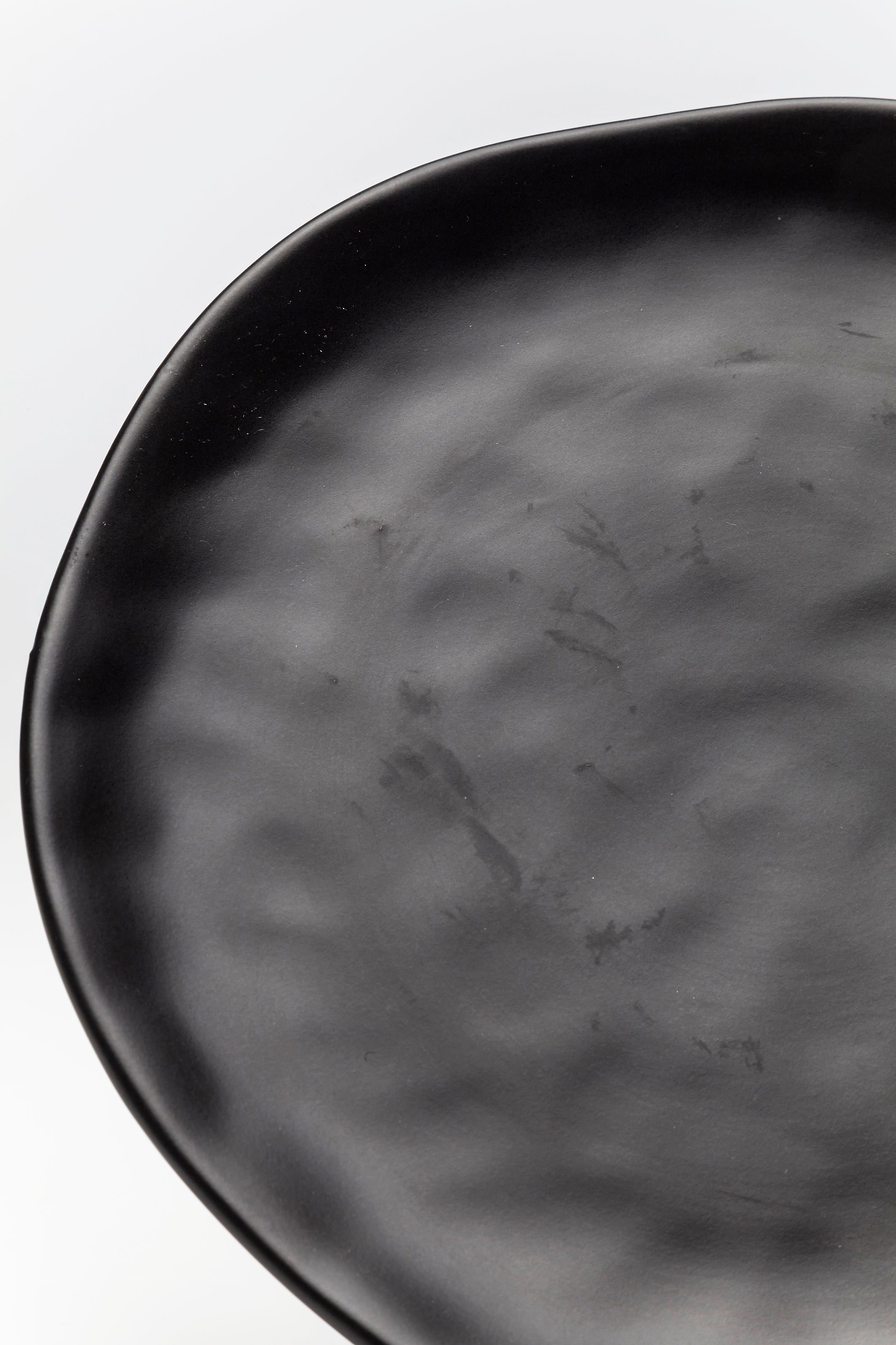 Eine Nahaufnahme des Tellers Organic Black Ø26 cm, ein mattschwarzer, asymmetrischer Steingut-Teller mit subtilen Glanzflecken und welliger Textur, der seinen handgemachten Look vor einem weißen Hintergrund hervorhebt.