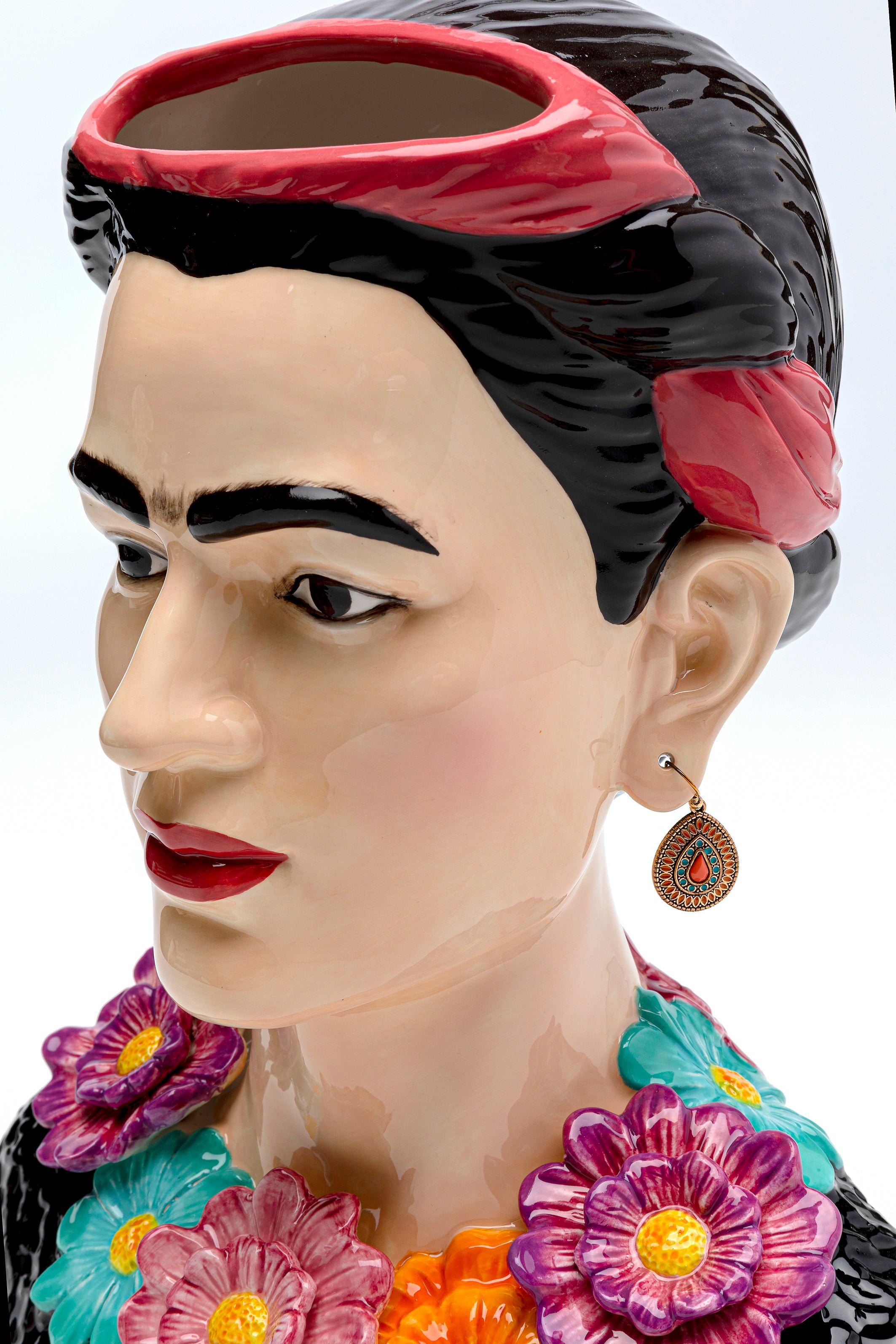 Die Deko Vase Style Muse Flowers ist eine farbenfrohe Keramikvase in Form eines Frauenkopfes mit dunklem Haar mit roter Schleife, markanten Augenbrauen, roten Lippen, einem Ohrring und einer Blumenkette. Die offene Oberseite ermöglicht kreative Blumenarrangements.