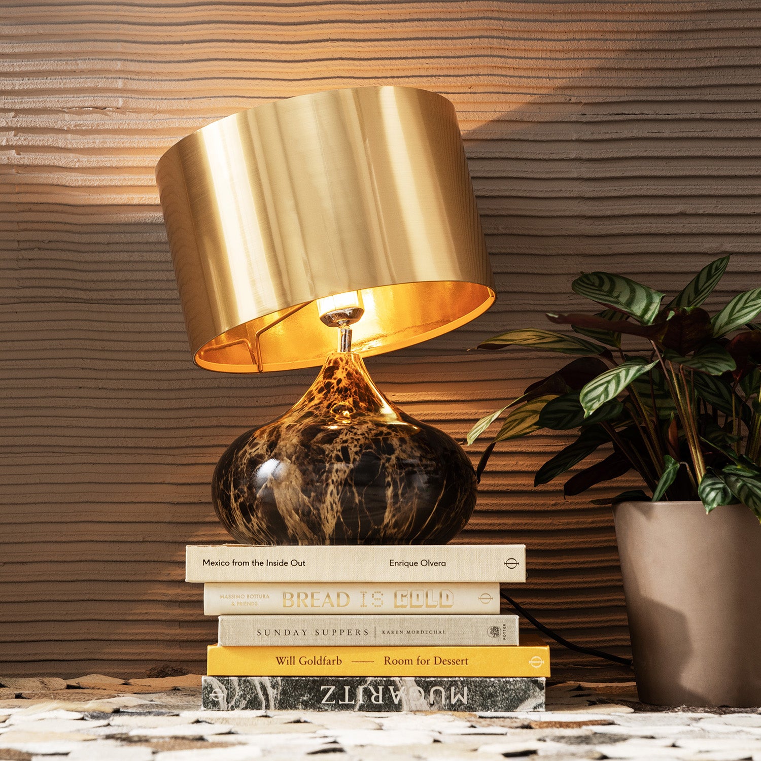 Die goldene Tischleuchte Mamo Deluxe mit Lampenschirm und Marmorsockel ruht auf einem Stapel Bücher neben einer Topfpflanze, die auf einer strukturierten Oberfläche mit einer geriffelten Wand im Hintergrund steht.
