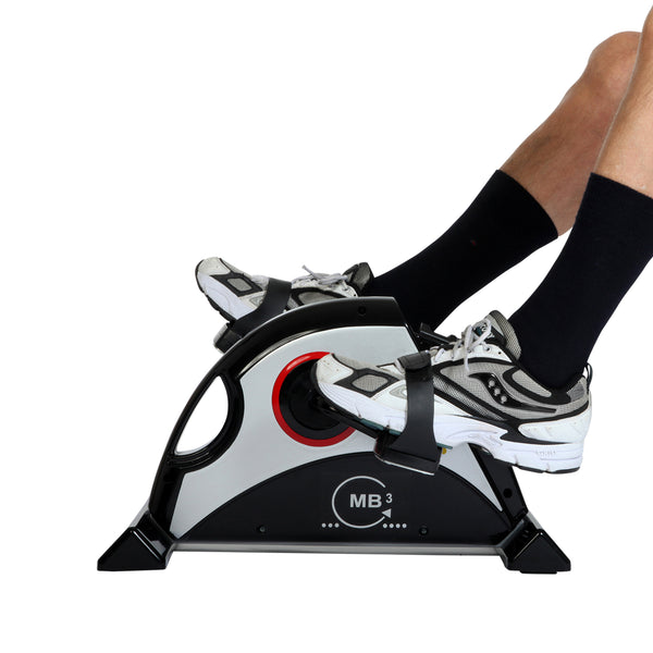 Person, die auf einem kompakten Heimtrainer in die Pedale tritt, mit schwarzen Socken und grauen Sportschuhen vor einem weißen Hintergrund.