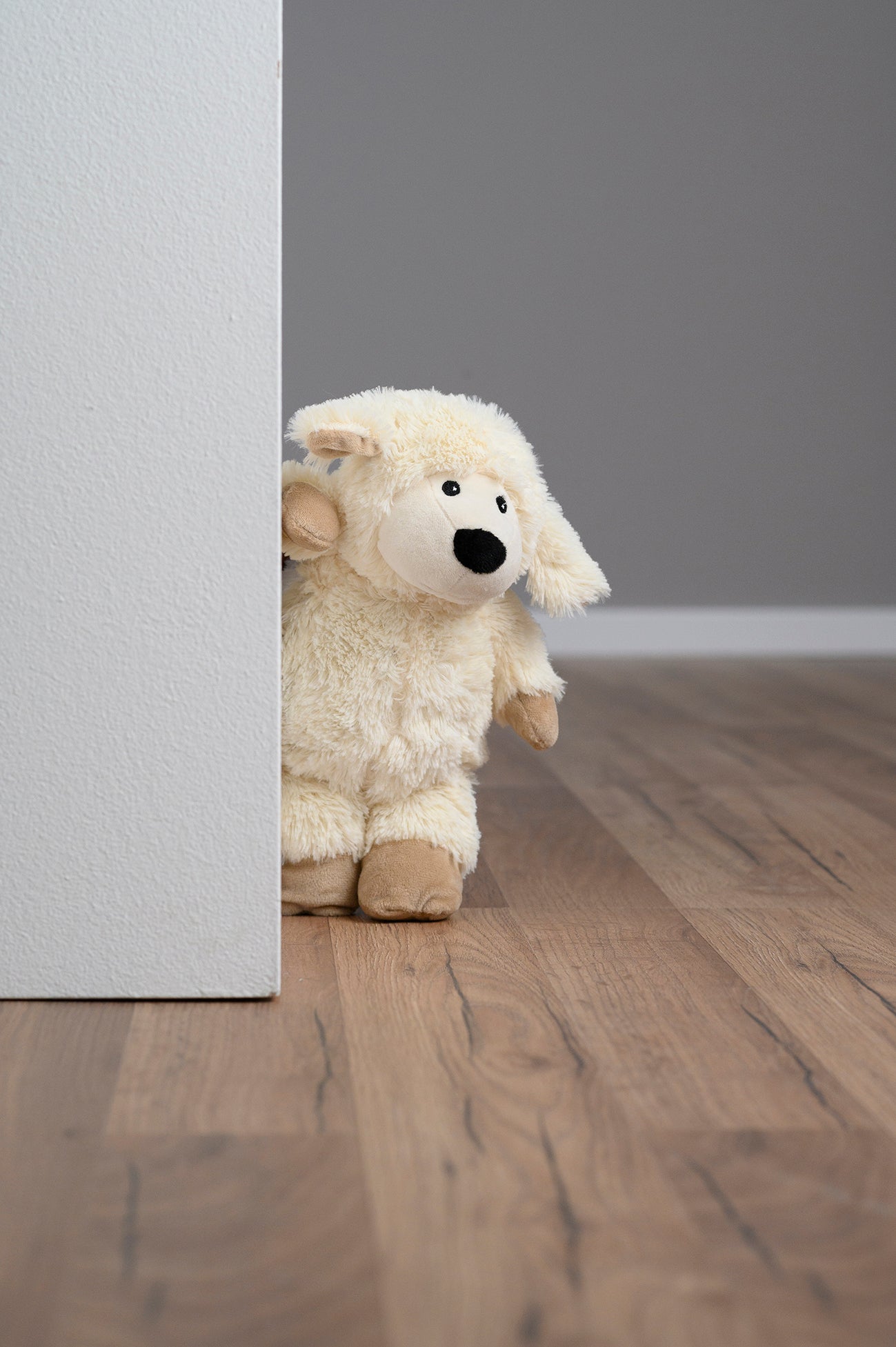 Hinter einer weißen Wand lugt ein Plüschschaf auf einen Holzboden hervor.