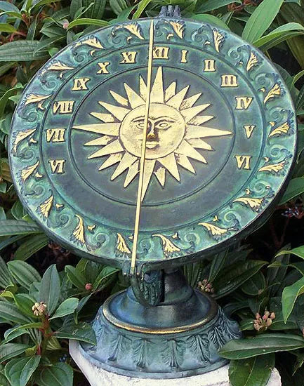 Eine dekorative Sonnenuhr mit römischen Ziffern und einem Sonnengesicht, umgeben von grünen Blättern.