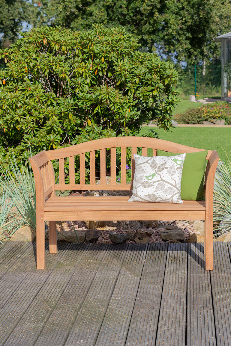Eine Holzbank mit zwei Kissen auf einer Terrasse in einem Garten mit grünen Büschen und Gras.