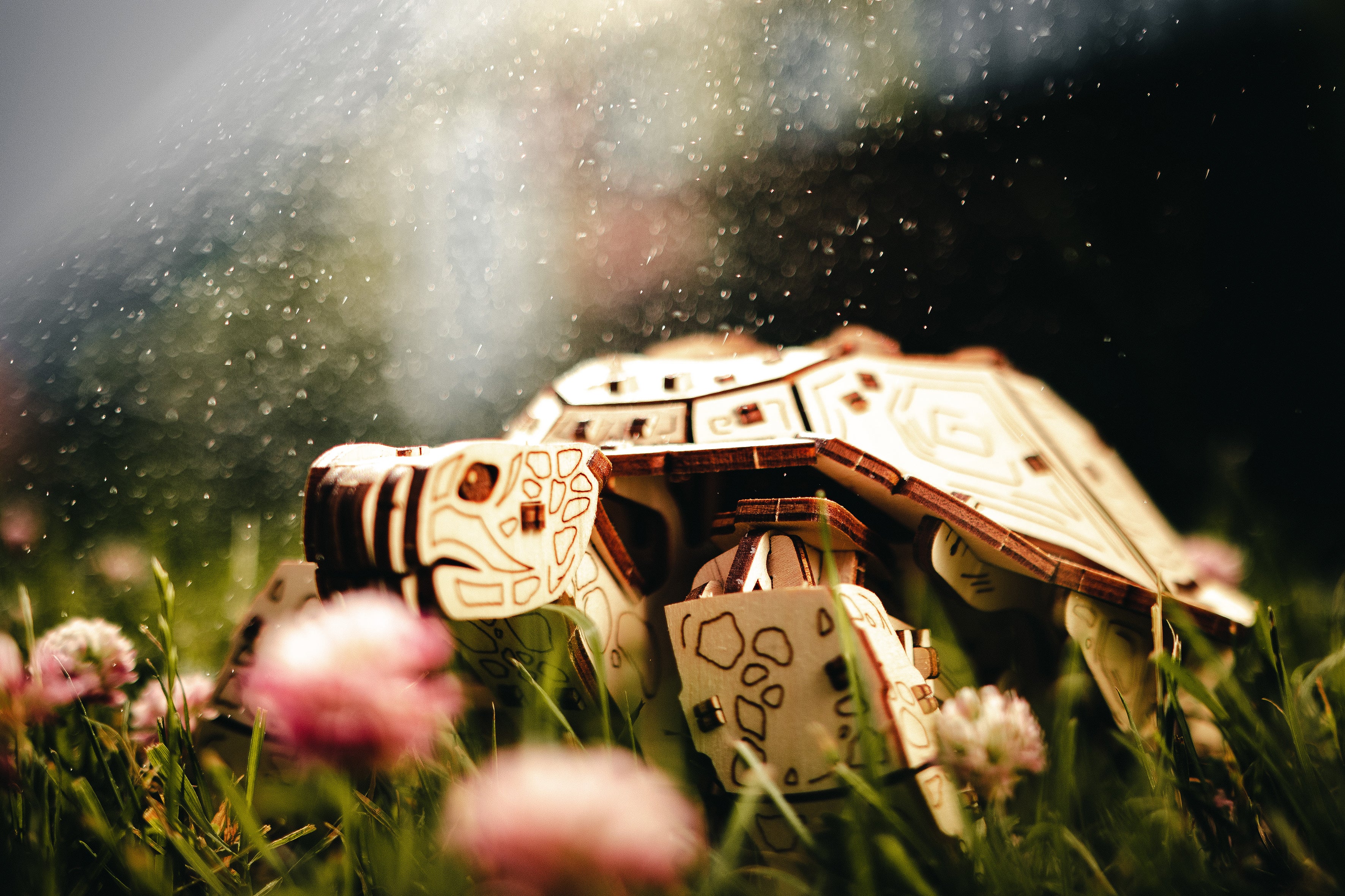 Eine Spielzeug-Roboterschildkröte sitzt im Gras und zwischen Kleeblüten, während Sonnenlicht durch Nebel oder Regen strömt.