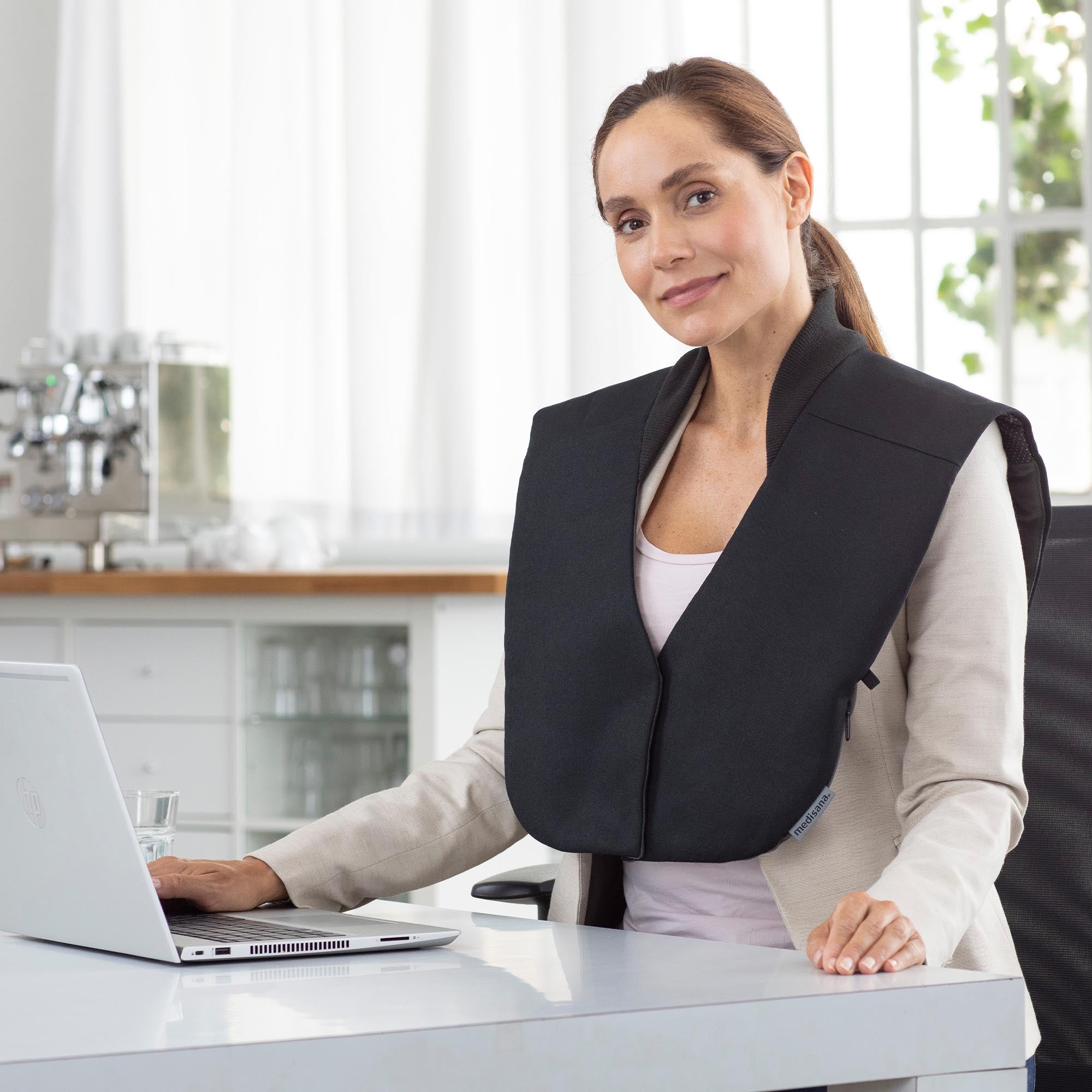 Frau trägt ein schwarzes Schultermassagegerät, während sie an einem hellen, modernen Schreibtisch an einem Laptop arbeitet.