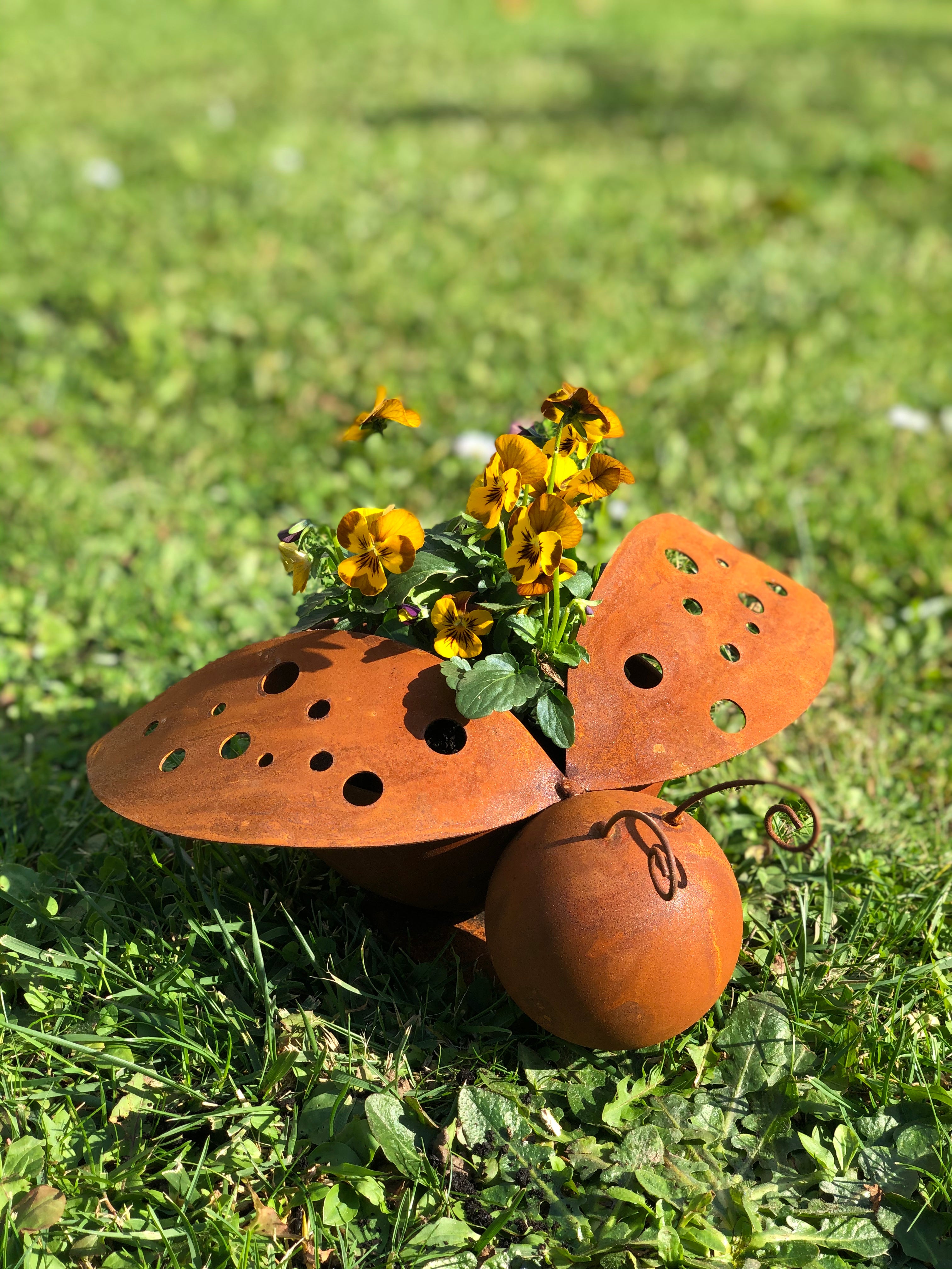 Schmetterlingsförmiger Blumentopf aus rostigem Metall mit gelben Blüten, der im Sonnenlicht auf grünem Gras steht.
