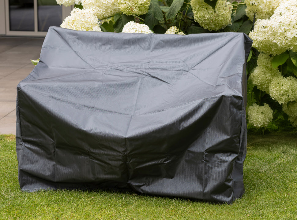Eine schwarze Gartenmöbelabdeckung auf einem großen Möbelstück, mit weißen Blumen und Grün im Hintergrund.