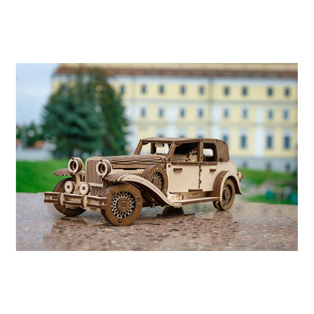 Holzmodell eines Oldtimers im Freien auf einer Steinfläche mit einem unscharfen Gebäude im Hintergrund.
