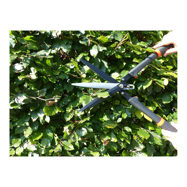 Hände, die mit einer Gartenschere mit schwarzem Griff eine belaubte Hecke stutzen.