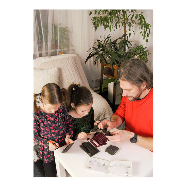 Ein Mann und zwei Mädchen bauen gemeinsam an einem weißen Tisch in einem gemütlichen, mit Pflanzen gefüllten Raum einen Modellbausatz zusammen.