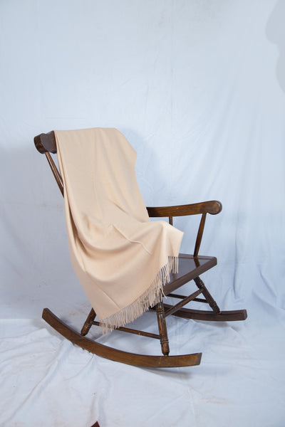 Ein hölzerner Schaukelstuhl, über dessen Rückenlehne eine beigefarbene Fransendecke drapiert ist, steht vor einem weißen Hintergrund.