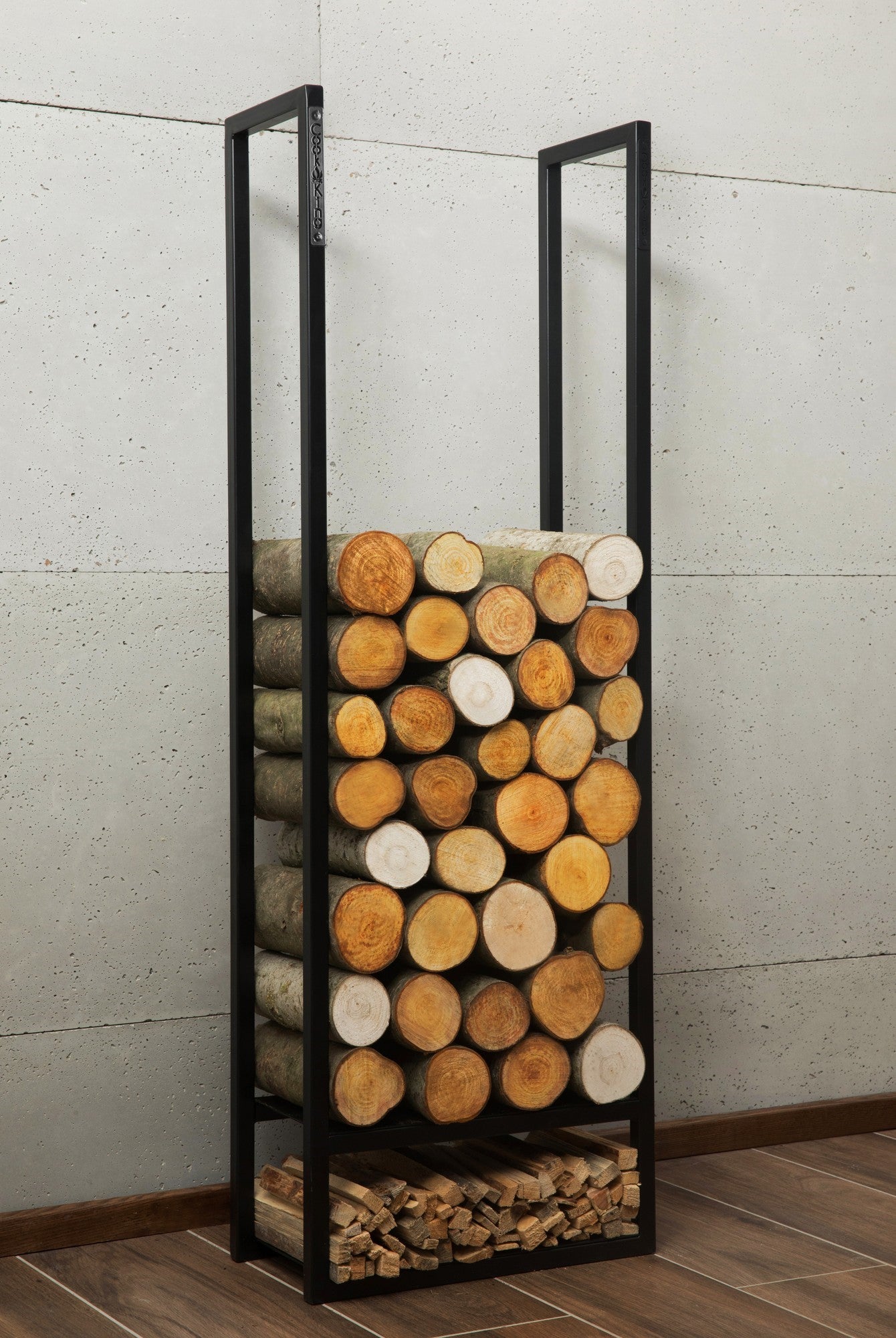 Ein hohes schwarzes Metallgestell mit gestapelten Holzscheiten und Anzündholz an einer Betonwand.