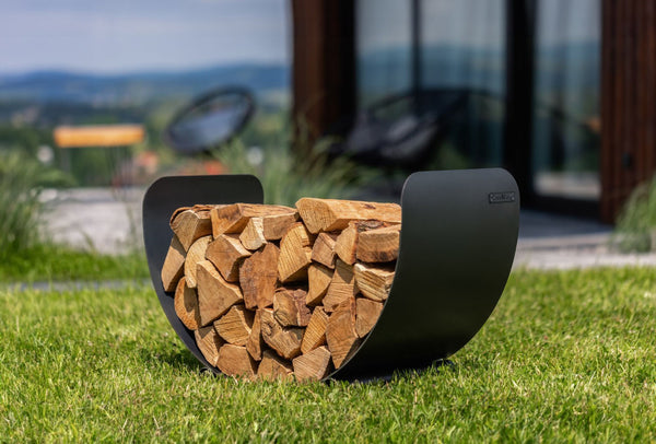 Ein gebogenes Metallgestell mit gespaltenen Holzscheiten liegt auf grünem Gras vor einem modernen Gebäude.