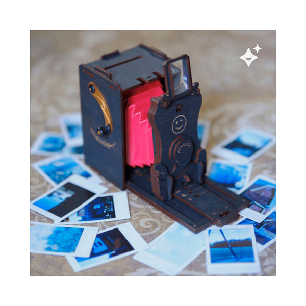 Ein hölzernes Kameramodell im Vintage-Stil mit rotem Balg, umgeben von verstreuten Sofortbildern.