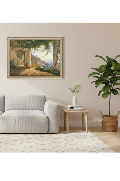 Die Edition zeigt eine mit Weinreben bewachsene Terrasse bietet einen Blick auf eine Küstenstadt und das Meer, eingerahmt von Säulen und üppigem Grün. Die ist in einem Wohnraum mit Sofa und Pflanze über den Sofa aufgehängt. 