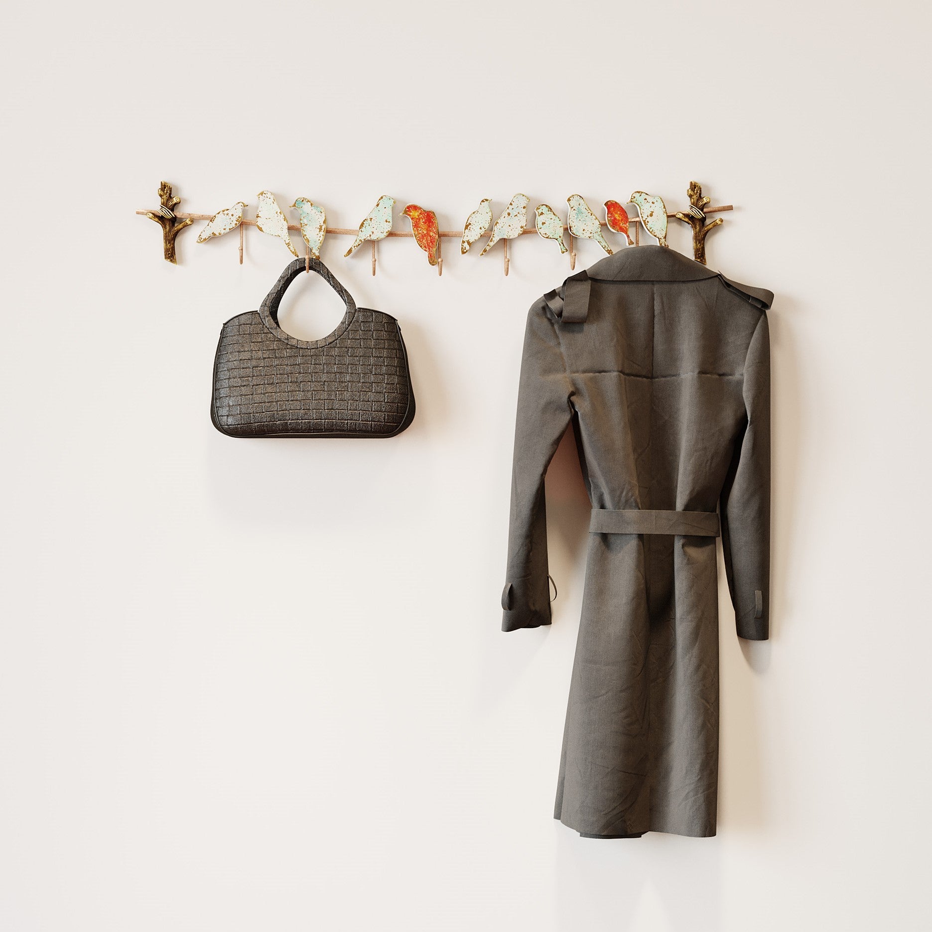 Eine graue Handtasche und ein dunkelgrauer Trenchcoat hängen an der Wandgarderobe Bird Party, einem dekorativen Wandhaken in Form eines Astes mit bunten Vögeln, der einer schlichten hellen Wand Charme verleiht.