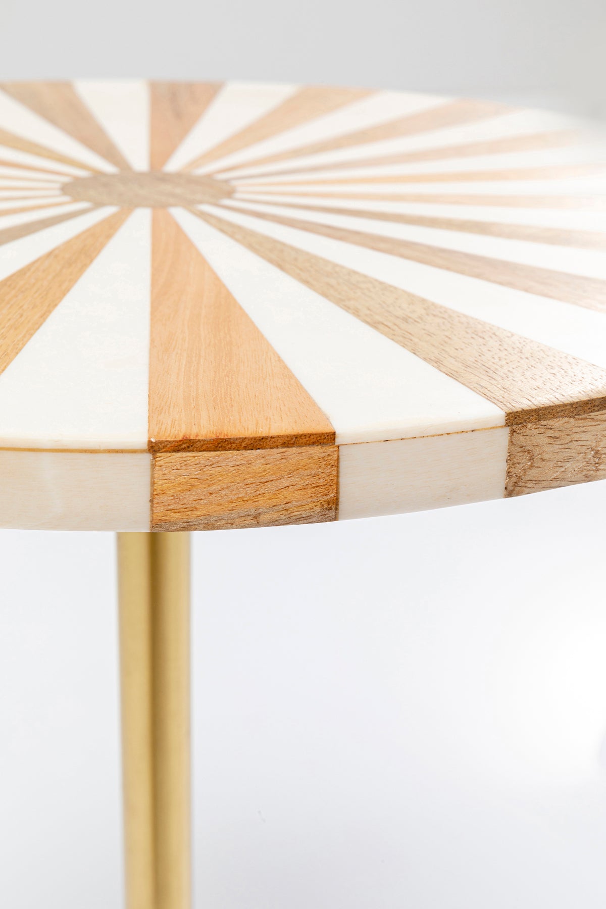 Eine Nahaufnahme des Beistelltischs Domero Cirque Gold Weiß Ø40cm zeigt sein Sonnenstrahlenmuster mit abwechselnden Holz- und Weißsegmenten, getragen von einem schlanken, goldbeschichteten Bein.