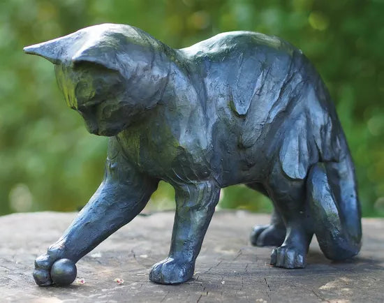 Die bronzene Gartenskulptur „Verspielte Katze mit Ball“ zeigt vor einer grünen Kulisse eine verspielte Katze, die mit ihrer Pfote nach einem kleinen Ball greift.
