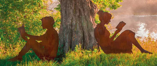 Silhouetten zweier Personen aus dem Gartenfiguren-Set „Lesendes Paar“ sitzen Rücken an Rücken lesend an einem Seeufer.