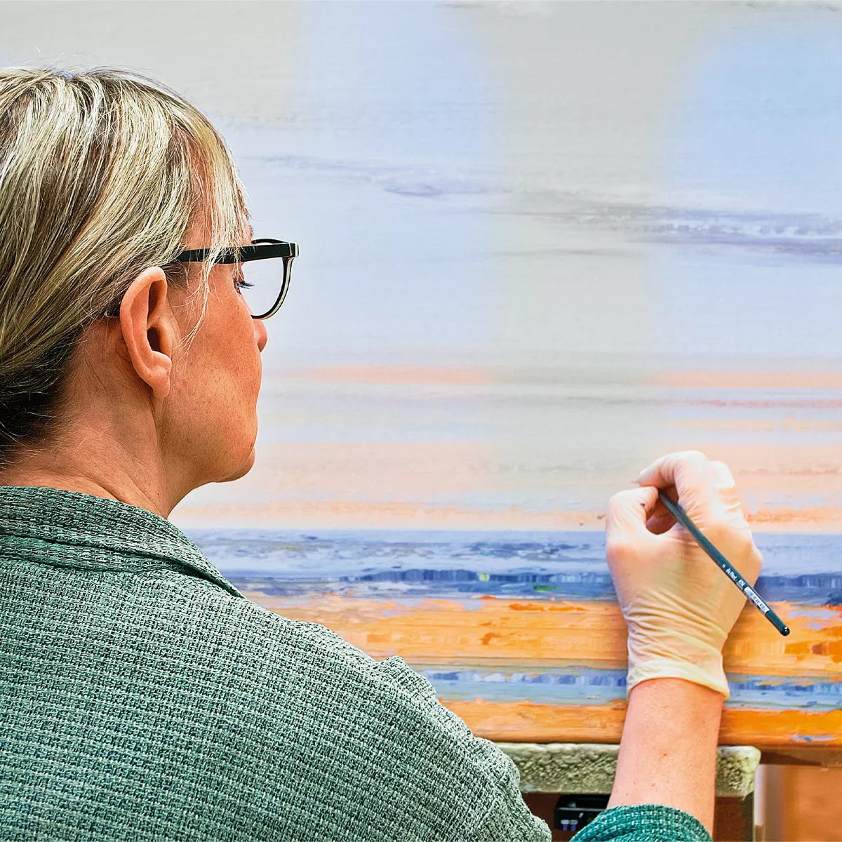 Eine Person mit Brille und Handschuhen malt „Leises Meeresrauschen – Jubiläums-Edition“ (2022, serielles Unikat) von Anja Struck auf eine Leinwand. Das gerahmte Kunstwerk zeigt eine farbenfrohe Landschaft.