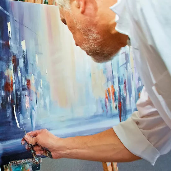 Ein Künstler malt eine lebendige Stadtlandschaft, wie sie in Robert Hettichs gerahmtem Werk „Golden Day – Jubiläums-Edition“ (2022, serielles Unikat) zu sehen ist, und hält dabei mit einem Spachtel jedes Detail fest.