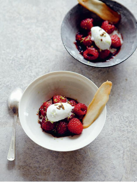 Zwei Schalen mit Himbeeren mit Sahne, einem knusprigen Keks und gehackten Nüssen, mit einem Löffel neben der einen Schale.