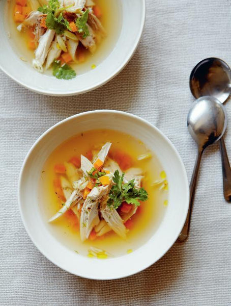 Zwei Schalen Hühnersuppe mit Gemüse und Kräutern, daneben zwei Löffel auf einem hellen Tischtuch.