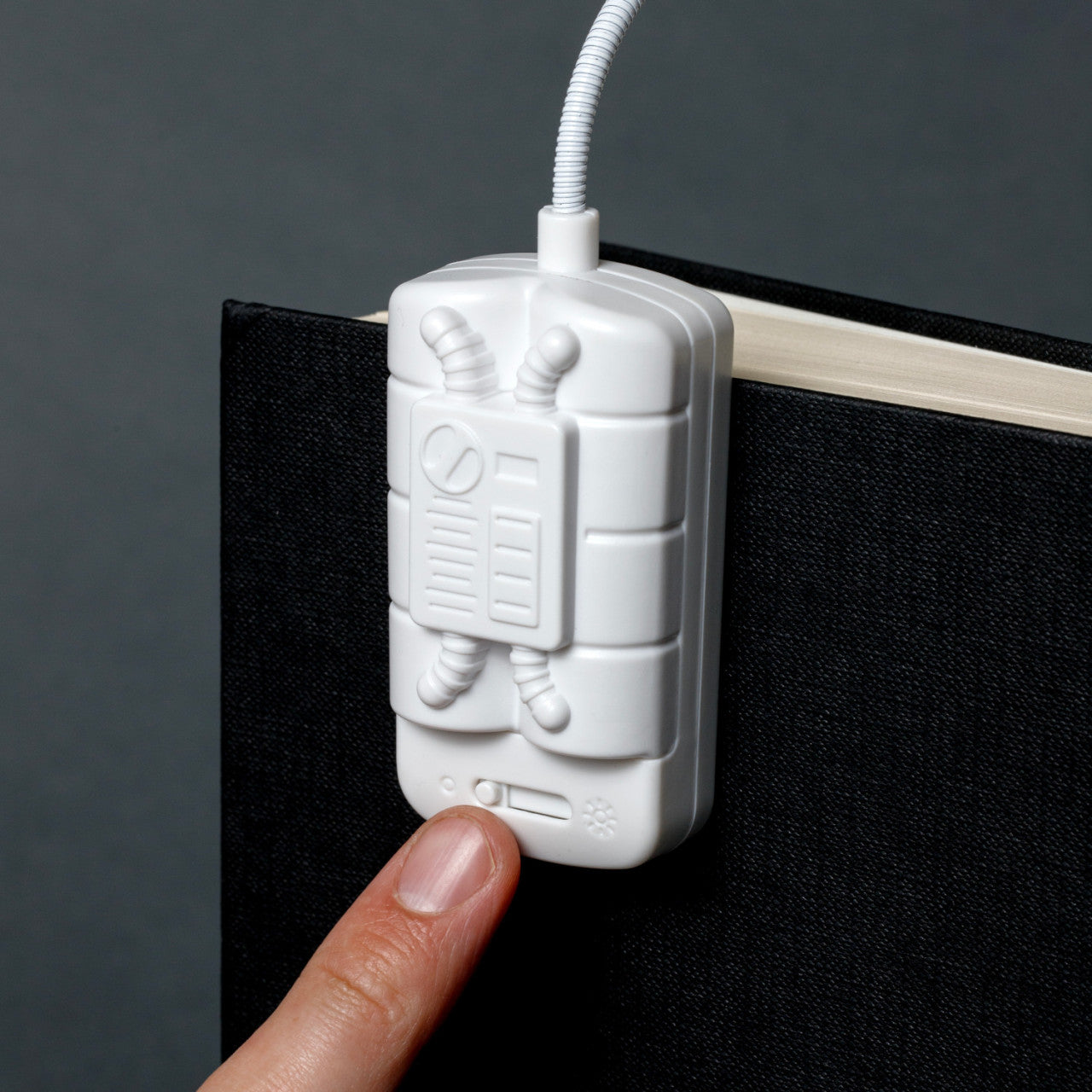 Eine Hand drückt auf die weiße Leselampe Spaceman – 2 LEDs, geformt wie ein Zünder, befestigt am Deckel eines geschlossenen schwarzen Buches.