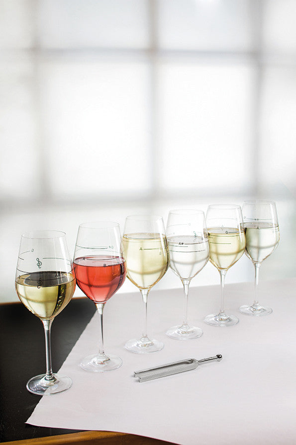 Auf einem weißen Tisch vor einem hellen Fenster stehen sechs Weingläser mit musikalischen Tönen, jedes mit einem anderen Wein.
