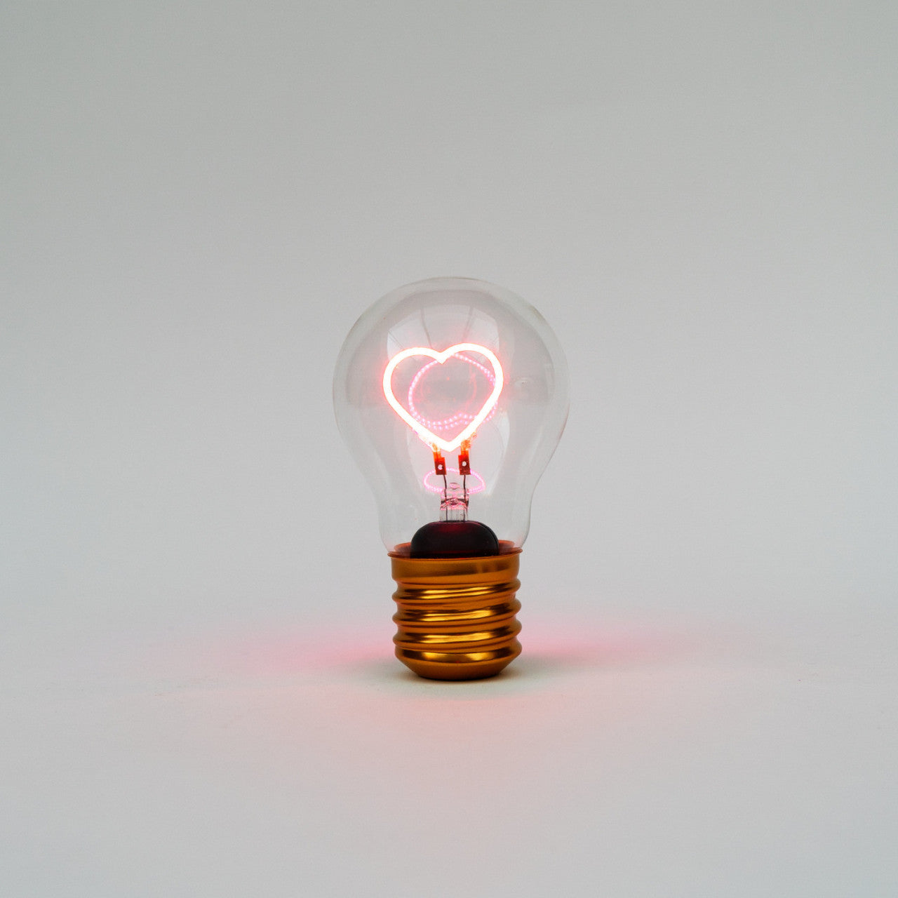 Die Kabellose LED-Glühbirne - Herz verfügt über einen leuchtenden rosa herzförmigen Glühfaden vor einem schlichten weißen Hintergrund.